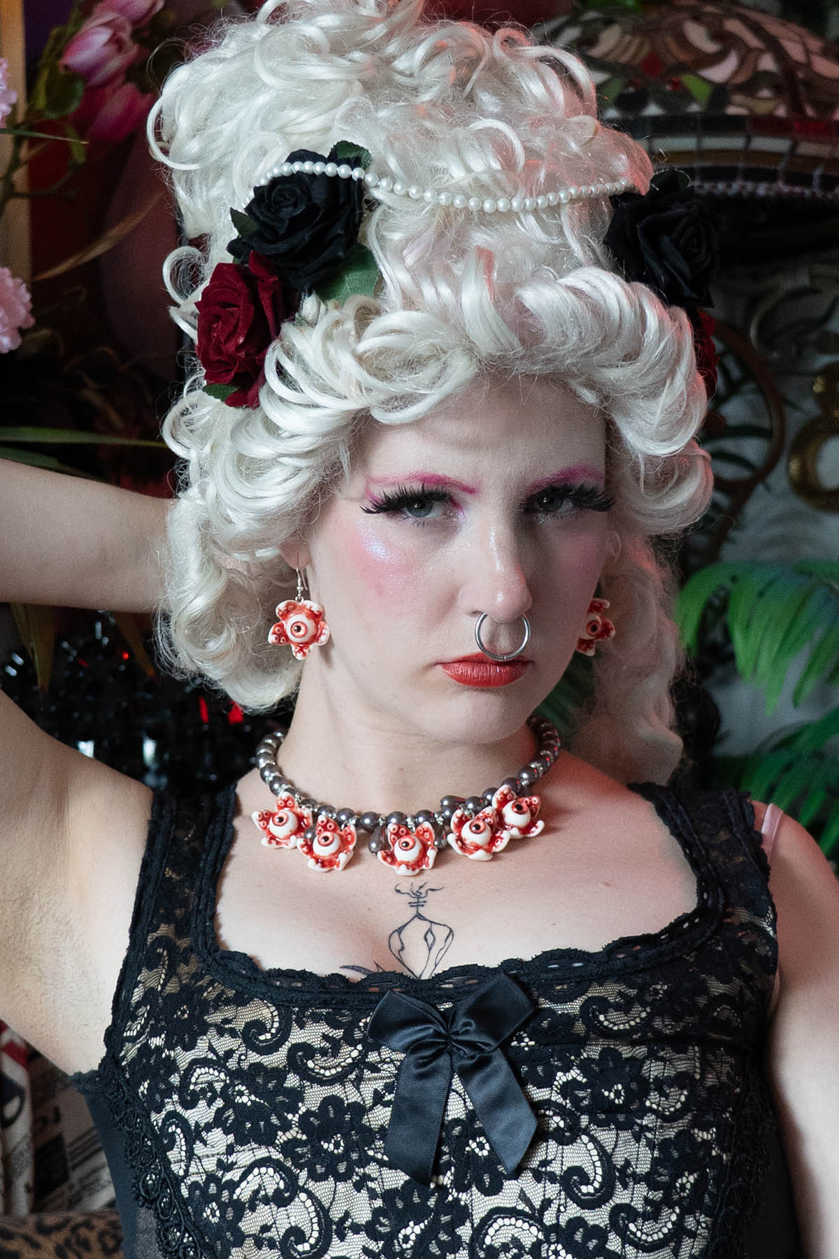 A person with white renaissance hair and makeup wearing a black lace top and a necklace with black glass pearls and resin eye ball pendants.