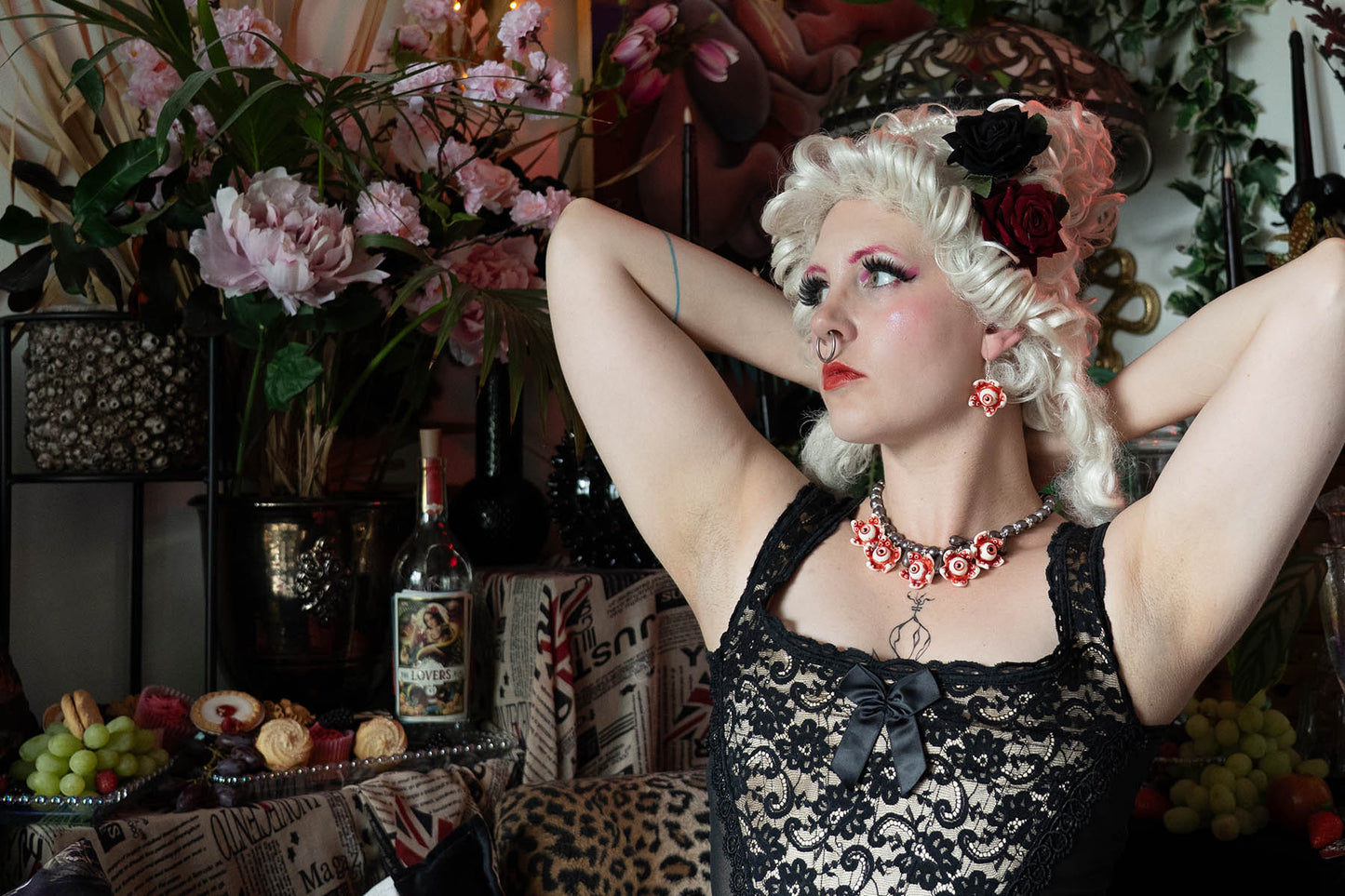 Woman in a lace dress with floral decorations and a pearl necklace with eyeballs, posing in a room with flowers and bottles.