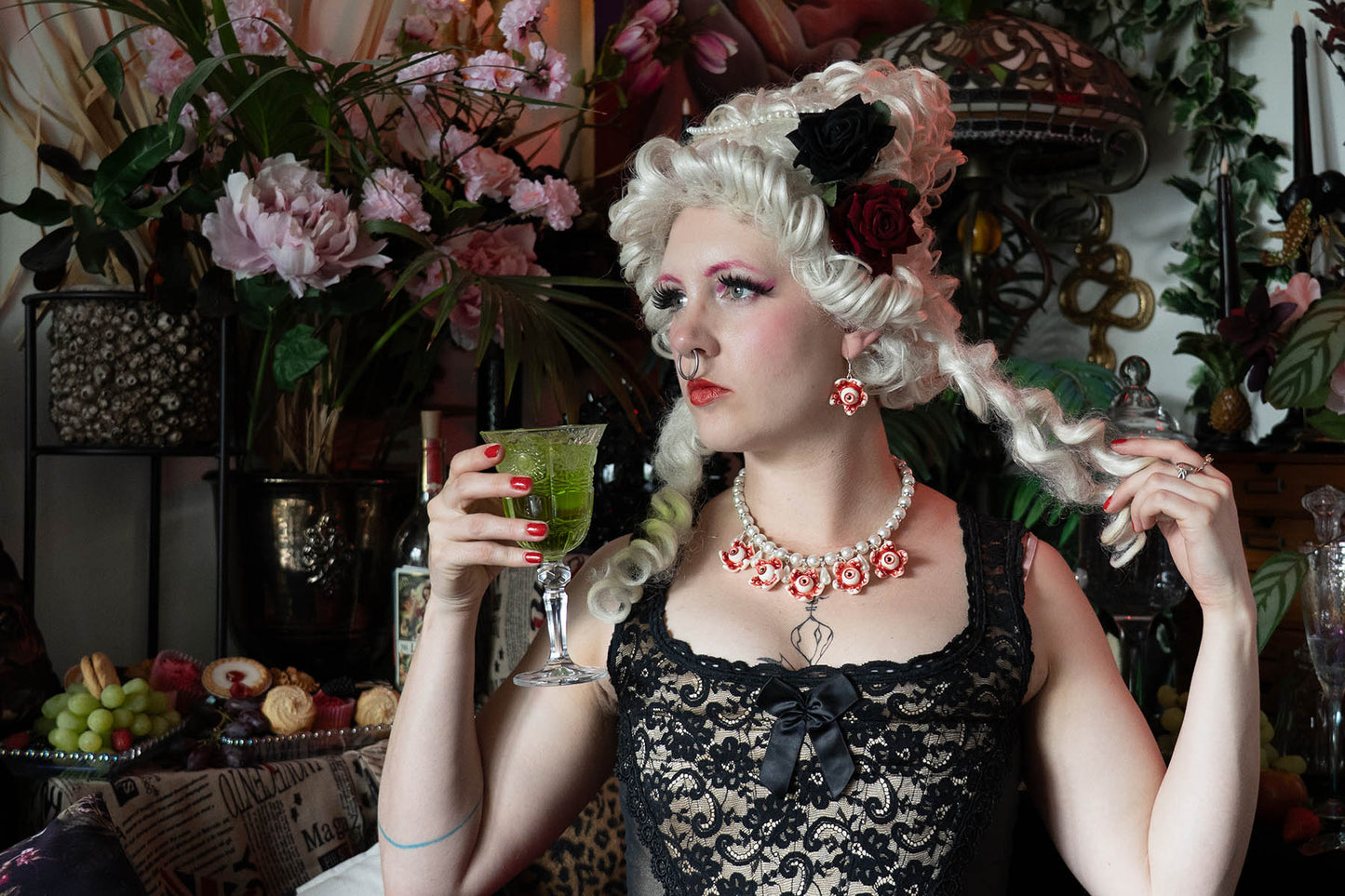 Person in vintage attire holding a drink in a decorative setting wearing a pearl necklace with eyeballs