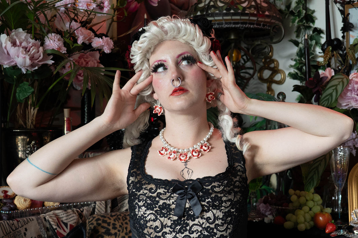 Person in a decorative setting with flowers, wearing a black lace dress and red and white jewelry.