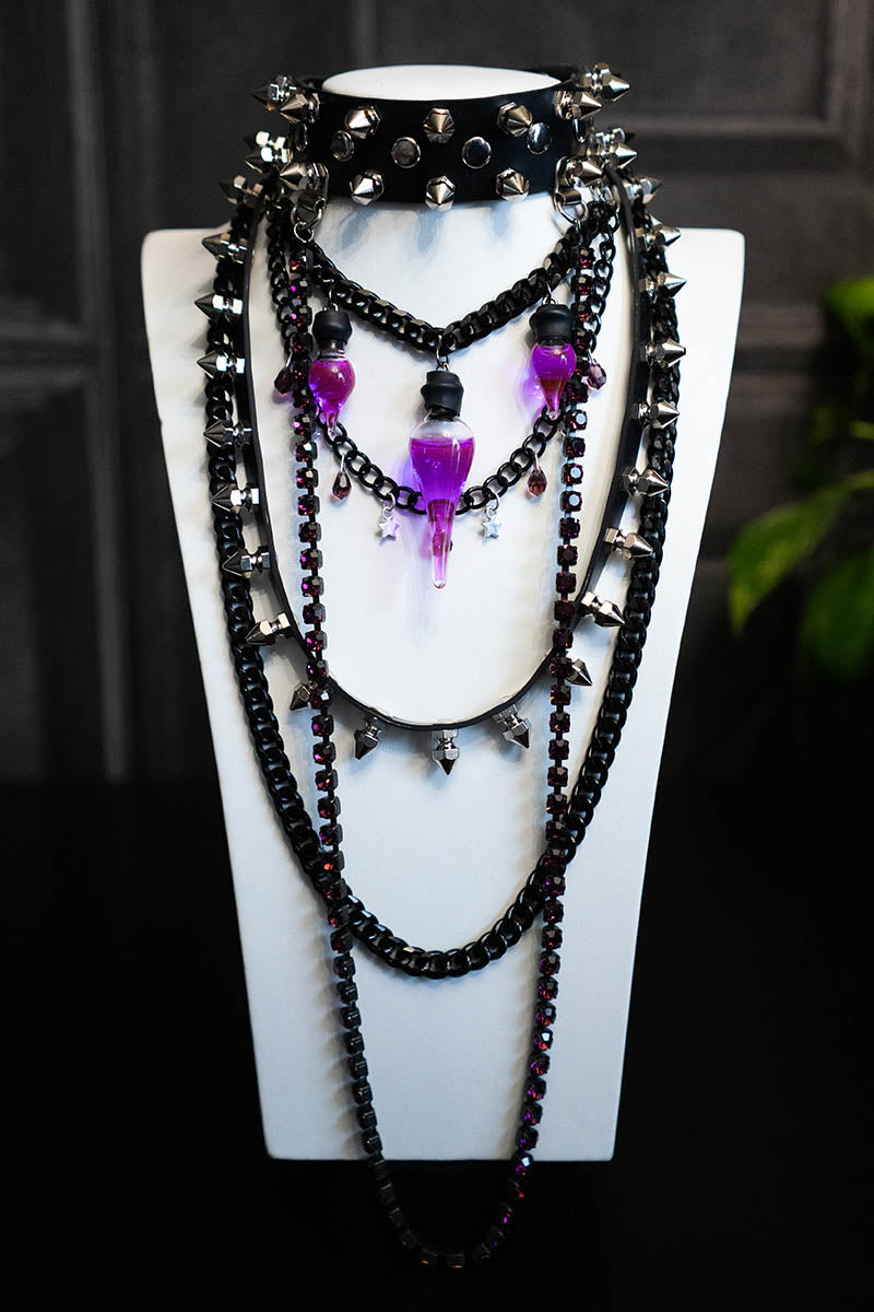 A black choker necklace with purple crystals, spikes, and a glass vial containing a purple liquid, displayed on a mannequin.