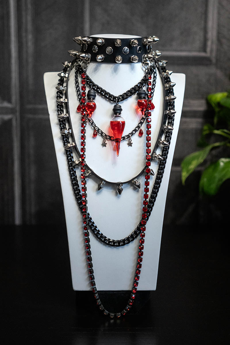 A layered-style choker necklace with black chains, red Preciosa crystals, and a glass bottle with red liquid, displayed on a mannequin.