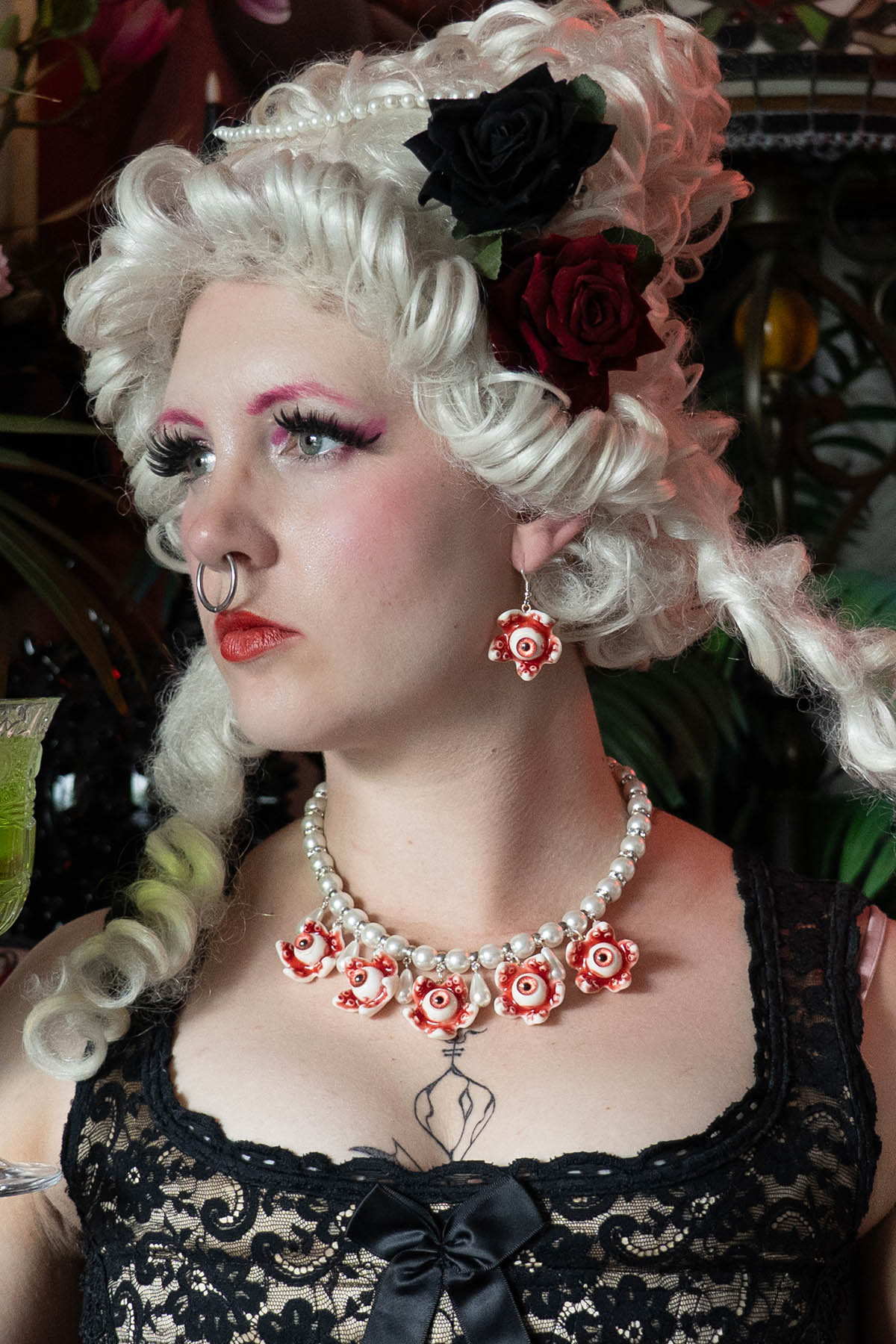 A person with white renaissance hair and makeup wearing a black lace top and a necklace with white glass pearls and resin eye ball pendants.