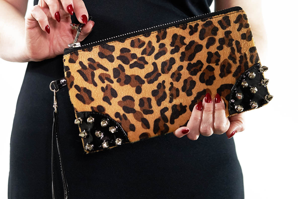 A leopard print clutch bag with black PVC spikes and a silver chunky zipper, being held by a person with red nail polish.