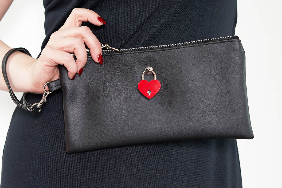 A black leatherette clutch bag with a red heart-shaped padlock on the front, held by a hand with red nails.