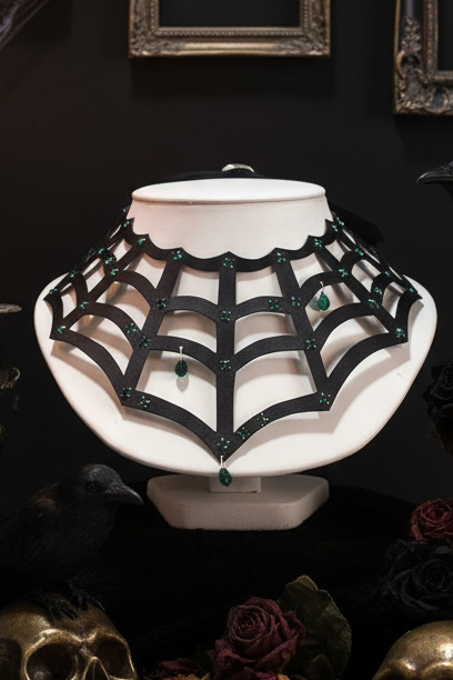 A gothic-style black spiderweb choker necklace with green gemstone accents, displayed on a white bust against a dark background with decorative skulls and roses.