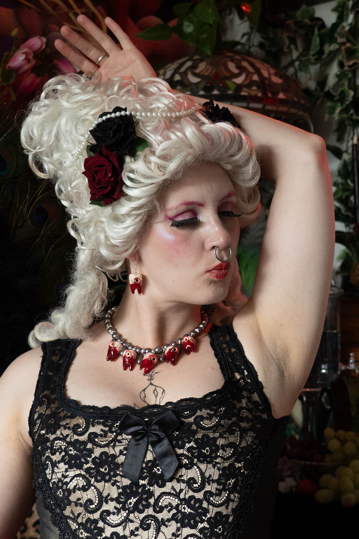 A person with white renaissance hair and makeup wearing a black lace top and a necklace with black glass pearls and resin teeth pendants.