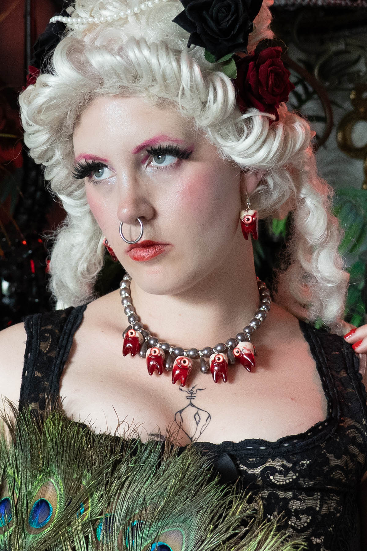 Person with styled renaissance white hair and dramatic makeup wearing a necklace with red teeth and black pearls