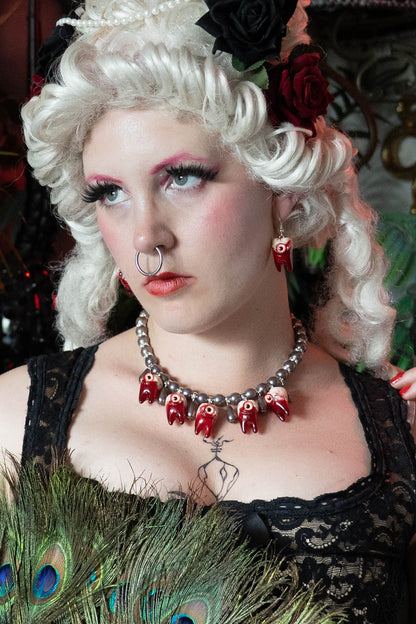 Person with styled renaissance white hair and dramatic makeup wearing a necklace with red teeth and black pearls