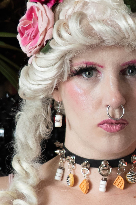 A person with a vintage hairstyle and makeup wearing silver milk bottle earrings, with a pink rose in their hair and decorative necklaces.