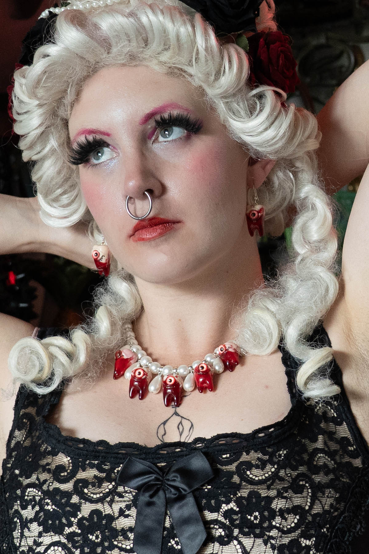 A person with white renaissance hair and makeup wearing a black lace top and a necklace with red glass pearls and gory teeth resin pendants.
