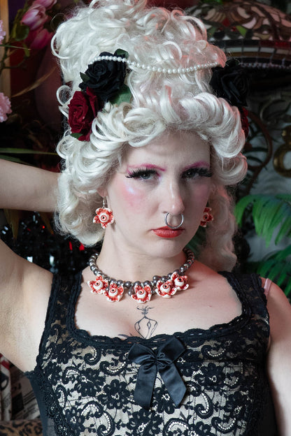 A person with white renaissance hair and makeup wearing a black lace top and a necklace with black glass pearls and resin eye ball pendants.