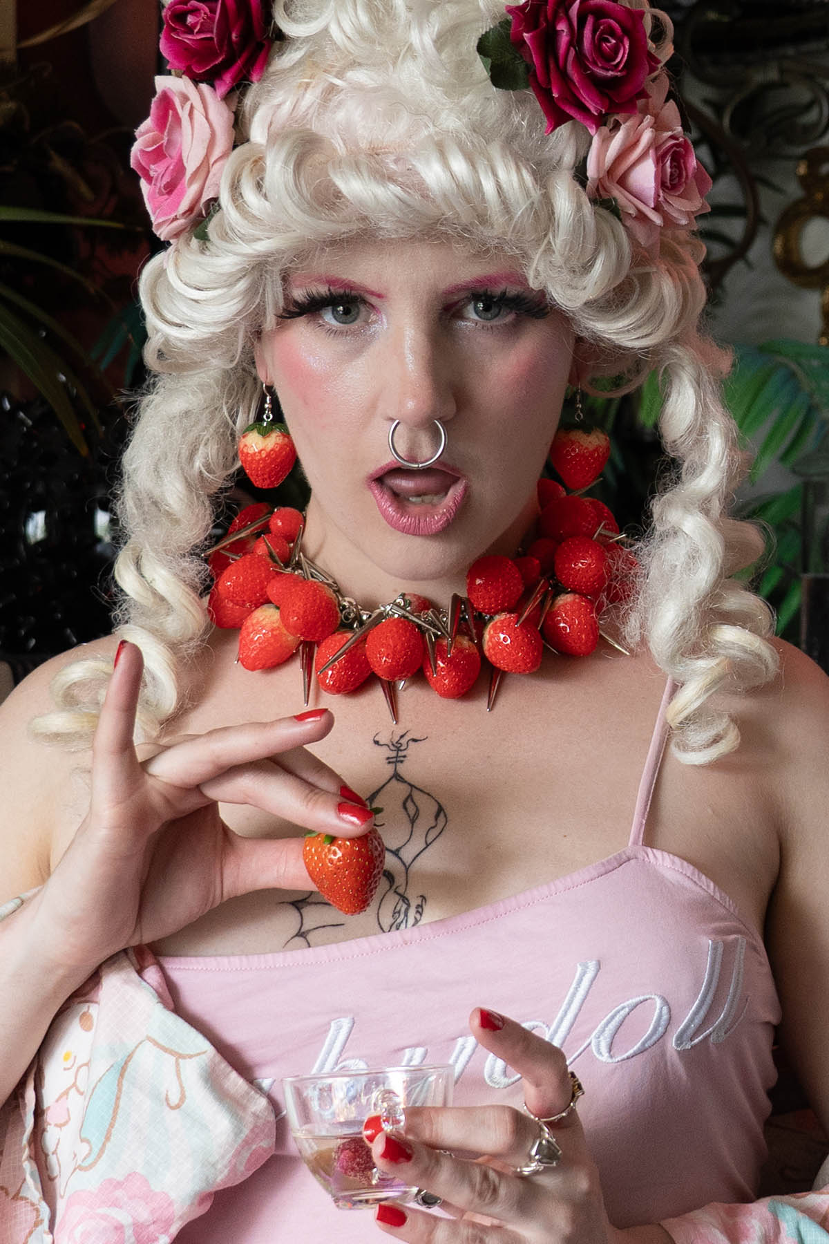 Person with white renaissance wig and floral headpiece wearing red strawberry-themed jewelry.