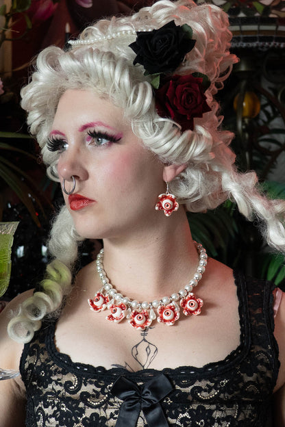 A person with white renaissance hair and makeup wearing a black lace top and a necklace with white glass pearls and resin eye ball pendants.