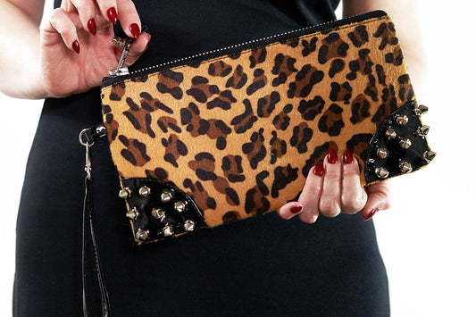 A leopard print clutch bag with black PVC spikes and a silver chunky zipper, being held by a person with red nail polish.