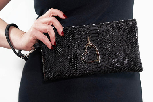 A black faux leather clutch bag with a heart-shaped carabiner and a removable strap, held by a person with red nail polish.